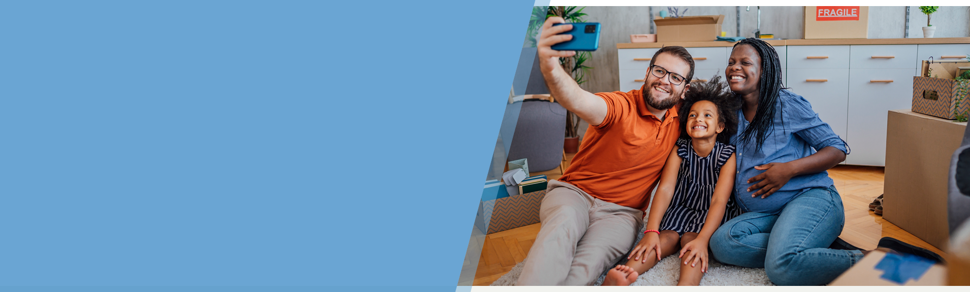 A family of three, including a man, a woman, and a child, sit on the floor taking a selfie. They are surrounded by moving boxes, suggesting they are in a new home. The left side of the image is a solid blue background.